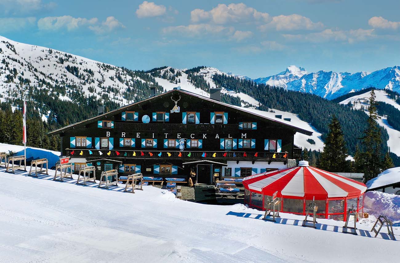 Die Breiteckalm im Winter: dunkles Holz, weiße Fensterläden und ein rot-weißer Sonnenschirm, dahinter die verschneiten Berge der Schmittenhöhe.